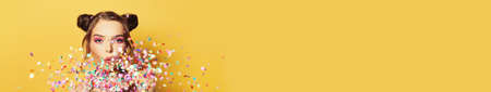 Beautiful woman blowing colorful confetti on yellow banner backgroundの写真素材