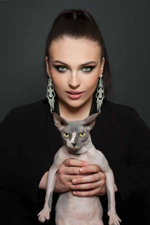 Fashion portrait of perfect elegant woman in diamond earrings with sphynx cat on blackの写真素材