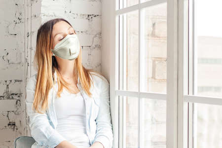 Woman in medical face mask near window at homeの写真素材