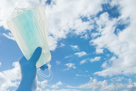 Hand in doctor gloves holding protective mask against sky clouds background, safety and virus protection conceptの写真素材