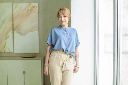 Cute smiling senior woman in blue casual blouse standing near windowの写真素材