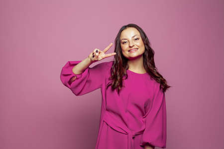 Pretty cheerful positive woman gesturing with fingers and smiling against pink wall studio...