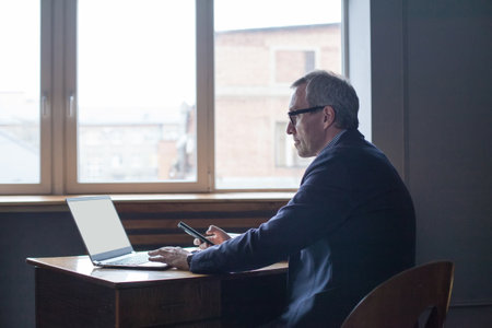 Elderly successful businessman in glasses sitting, working on laptop and holding mobile phone against window backgroundの写真素材