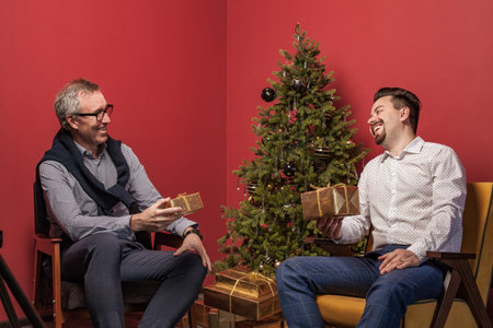 Elderly man and his son laughing and exchanging Christmas presents on red background and Christmas treeの写真素材