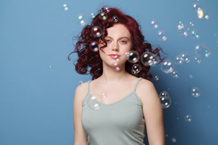 Beautiful woman with red curly hair and soap bubbles on blue studio wall backgroundの写真素材