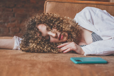 Beautiful woman with curly hair lies on the sofa with mobile phoneの写真素材