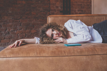Beautiful glamorous woman in white shirt with curly hair resting on the sofa with green mobile phone nearの写真素材