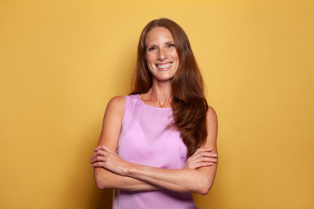 Portrait of pretty mid adult fashion model woman with long shiny red hair standing with crossed arms on vivid yellow backgroundの写真素材