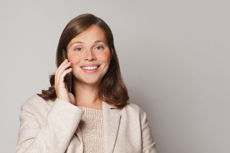 Attractive young woman talking on the phone and smiling while standing against grey studio wall backgroundの写真素材