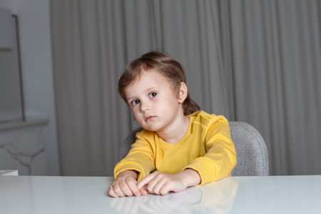 Pensive child boy with brown hair in yellow sweater sitting by the table, looking to the camera and pulling his handsの写真素材