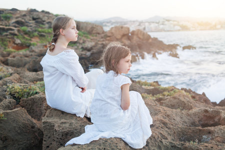 Lovely sisters in white clothes, kid and teenage girl sitting together near seaの写真素材