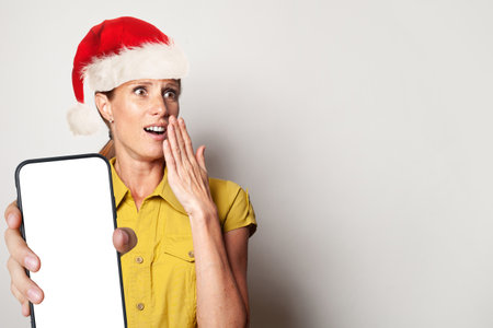 Excited surprised woman in Santa hat holding smartphone with empty blank display screen and present box on white backgroundの写真素材