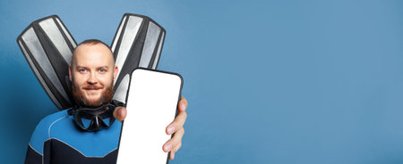 Happy diver diving man with flippers holding smartphone with empty white screen display for new Application on blue studio wall background. Mockup bannerの写真素材