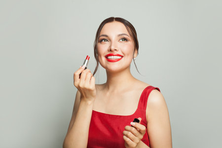 Pretty female beauty model is holding a red lipstick in one hand, She is wearing a red silky dressの写真素材