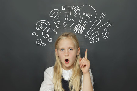 Excited child girl student with yellow light bulb and question marks on school blackboard background. Creativity, education and idea conceptの写真素材