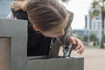 Cute child girl is drinking water from a fountain. Concept of refreshment and hydration, as the girl is taking a break from her day to quench her thirstの写真素材
