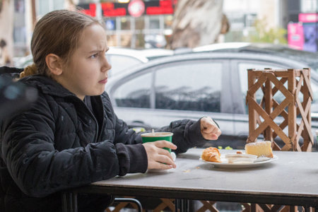 Tired young child girl drinking coffee and eating croissant in city street cafeの写真素材