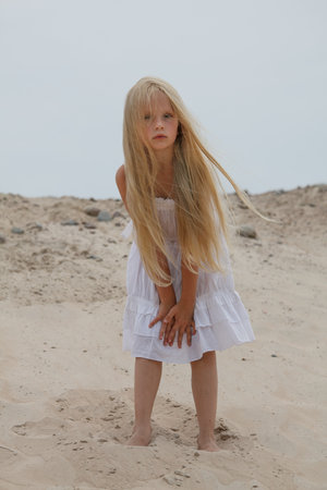 Beautiful young blond child girl in white dress standing on sand outdoorsの写真素材