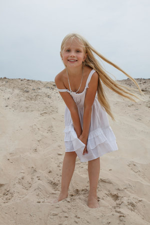 Joyful young blond child girl in white dress standing on sand outdoorsの写真素材