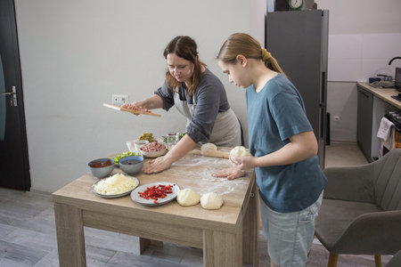 Little girl daughter and mother preparing dough for pizza and smiling while baking in kitchen at home, family togetherの写真素材