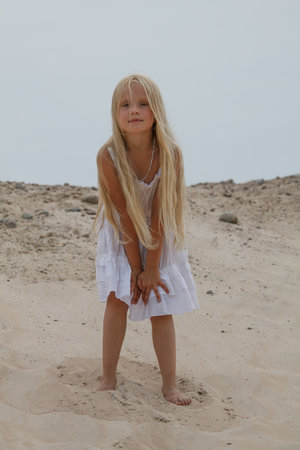 Portrait of young blond child girl in white dress standing on sand outdoorsの写真素材