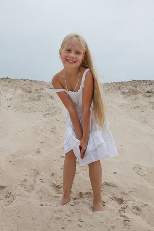 Smiling young blond child girl in white dress standing on sand outdoorsの写真素材