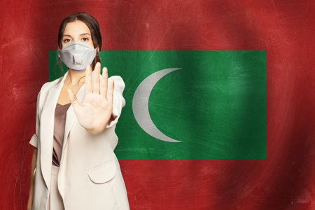 Confident woman in medical mask showing stop gesture open palm hand against flag of Maldives backgroundの写真素材