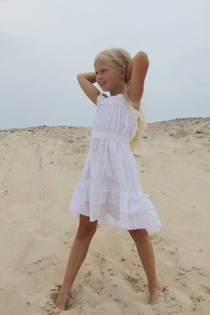 Cheerful young blond child girl in white dress standing on sand outdoorsの写真素材
