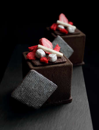 Dark chocolate textured cube dessert with strawberry, silver cookie decorations and red spongeの写真素材