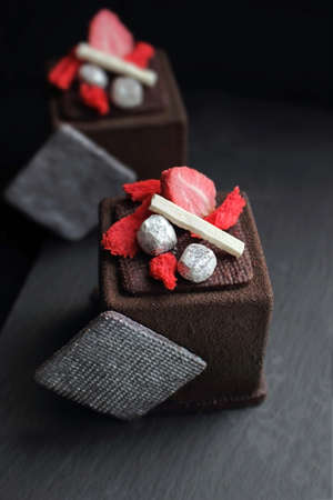 Dark chocolate textured cube dessert with strawberry, silver cookie decorations and red sponge on black backgroundの写真素材