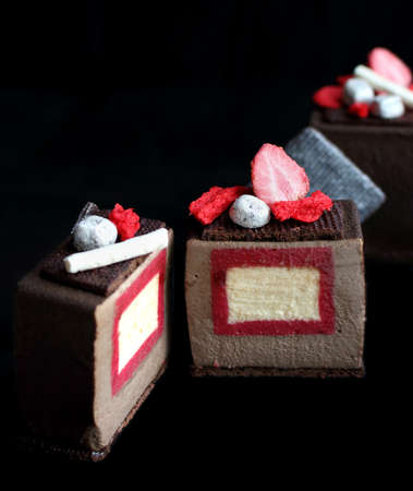 Sliced dark chocolate textured cube dessert with strawberry and vanilla insertion, silver cookie decorations and red sponge on black backgroundの写真素材