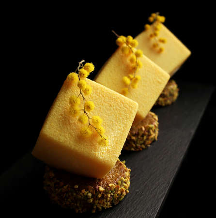 Yellow orange mousse textured desserts on pistachio sponge cake slices with mimosa flowersの写真素材