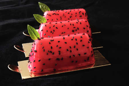 Red watermelon glazed desserts with black sesame seeds and basil leaves on golden coasters isolated on black backgroundの写真素材