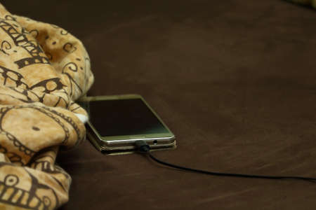 Silver mobile phone lying charging with cable on bed with blanketの写真素材