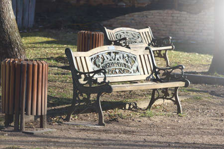 Wooden carved bench in park with trash bin standing in sunlightの写真素材