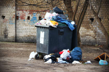 Trash container full of bags and clothes standing near tree in the yard