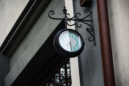 Old dirty round black outdoor clock with teal digits on black carved metal decorative holder hanging...