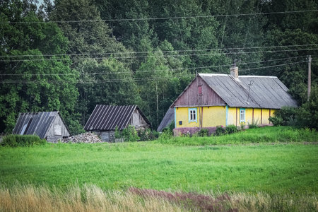 Small yellow farm house in typical surroundings of forest and grass meadow with visible field before harvest in Europeの写真素材