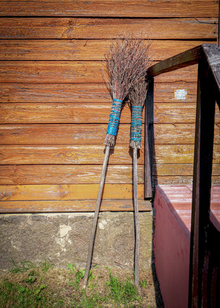 Two old brooms with blue rope holding all branches together standing up against the wall of wooden plank house with iron railing on sunny dayの写真素材