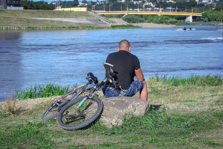 Man with bicycle sitting on o big stone looking at a river blue water and bike lying on green grass near byの写真素材