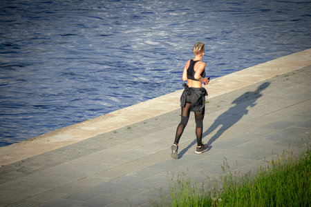 Young woman with short hair in dark sportwear running along a riverbank on a concrete path near...