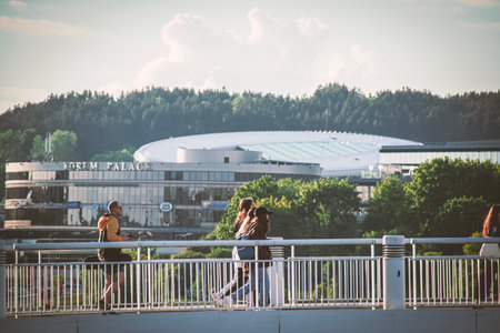 Vilnius, Lithuania 03 06 2022: Young people and men walking on a white bridge over river with green trees and office building on backgroundのeditorial素材