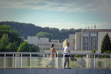 Vilnius, Lithuania 03 06 2022: Young woman an man walking on a white bridge with green trees and office building on backgroundのeditorial素材