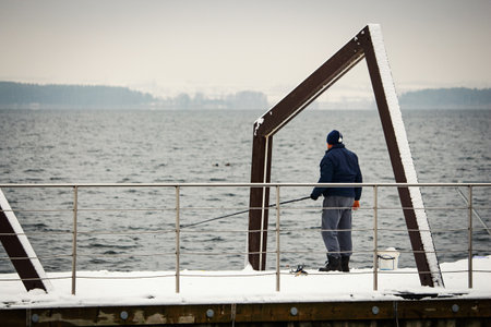 Man fishing in lake water waves under metal steel abstract architectural design arch on a fenced platform over lake with visible forest on opposite shoreの写真素材