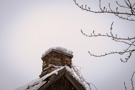 Snow covered old chimney and house roof top framed with leafless tree branches on a dull sky backgroundの写真素材
