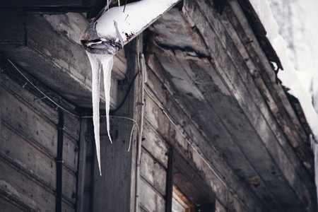 Steel broken drainpipe with icicles hanging on gray wooden building roof corner on winter dull day