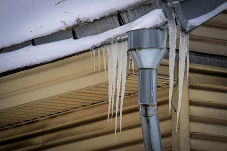 Steel drainpipe with icicles hanging on yellow building roof corner on winter dull dayの写真素材