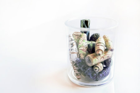 Handmade paper beads of brown, green, black colored floral patterns in plastic glass on white table side viewの写真素材