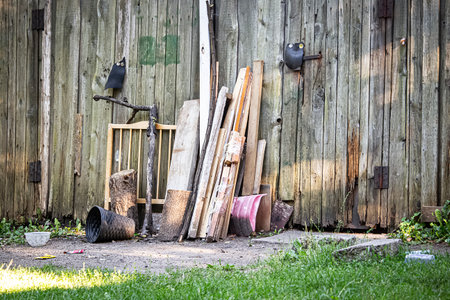 Random garbage like winter shovel, a trash bin, and wooden planks near a wooden garage wall on sunny...