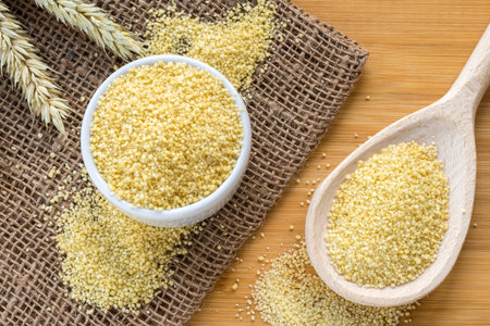 Couscous grains (durum wheat) in wooden spoon and ceramic bowl placed on the bamboo table, flat lay...